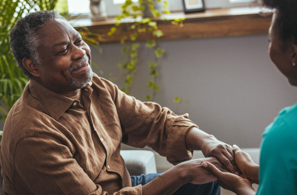 A senior with Parkinson's smiling while clasping hands with an out-of-frame caregiver while sitting near the window in senior living.