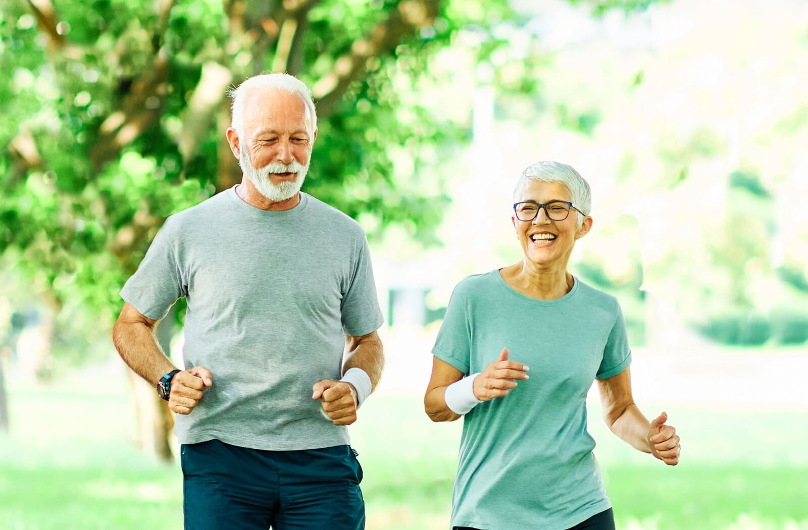 Two older people jog in the park with big smiles on their faces.