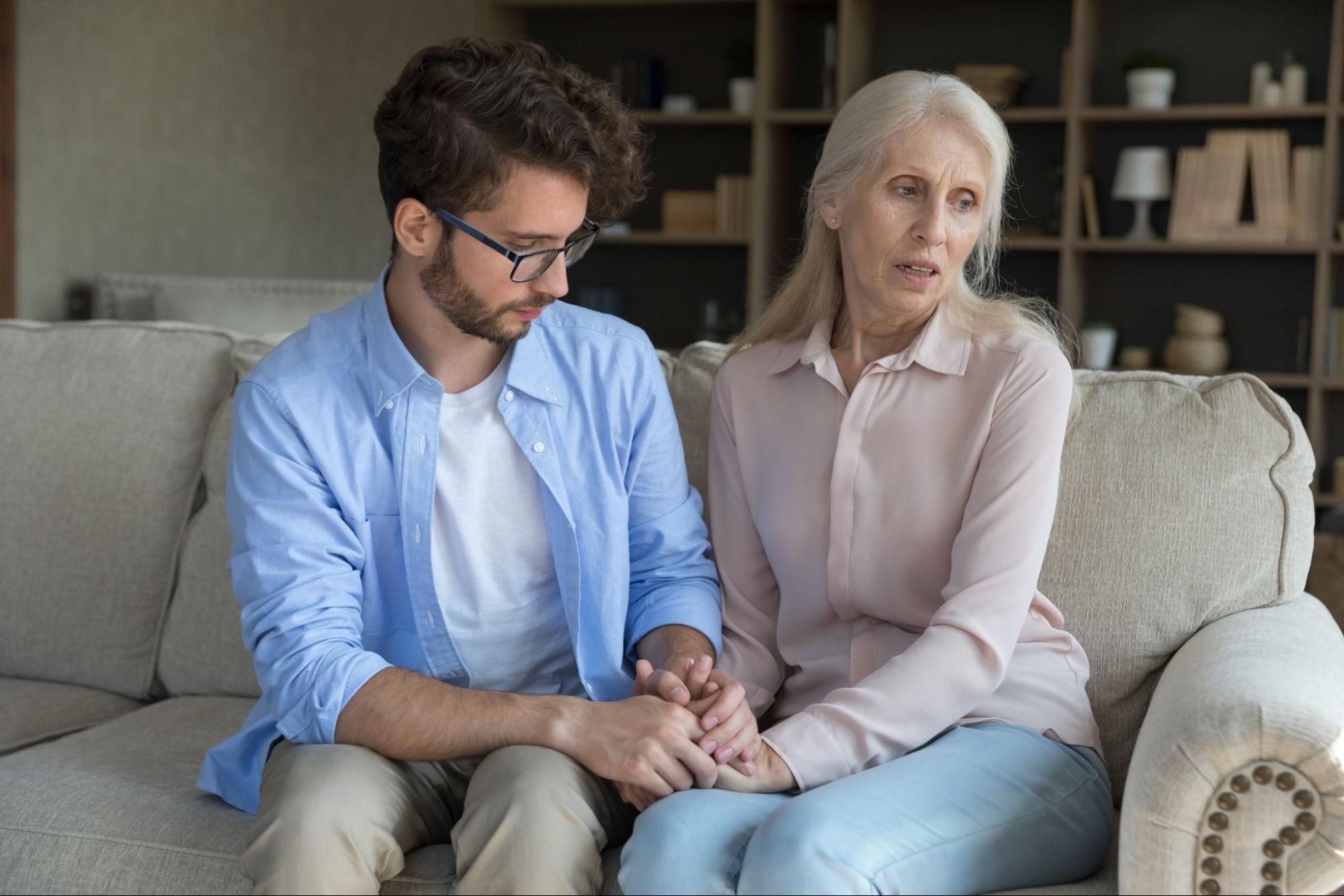 An adult child sitting next to their elderly parent holding their hands.