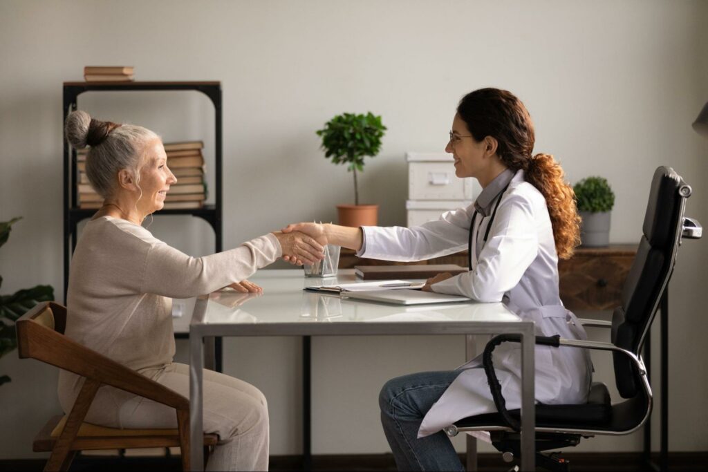 An elderly parent sitting in an office shaking a healthcare professional's hand.