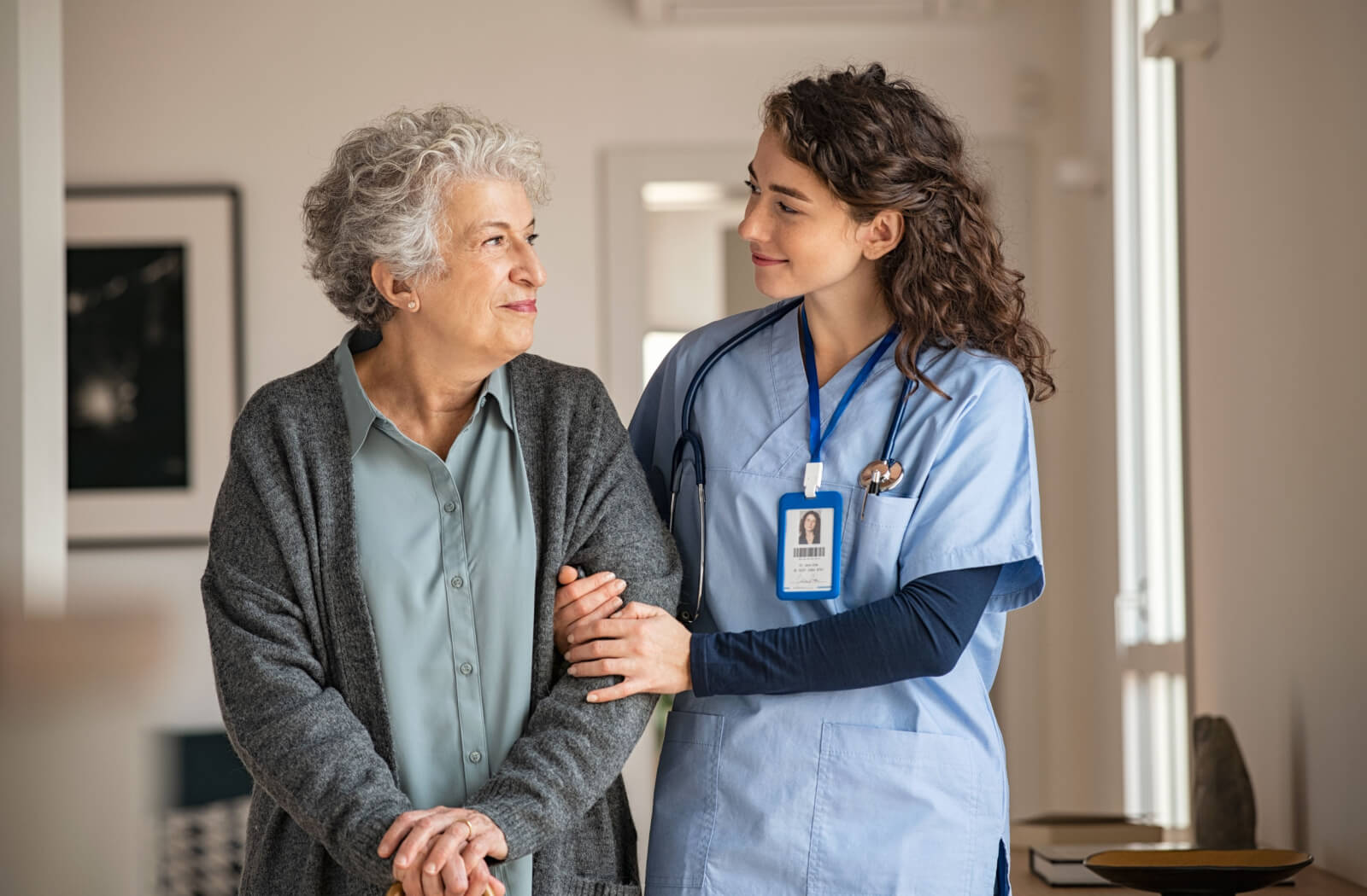 A young caregiver and a senior with Parkinson's walking arm-in-arm, looking at one another with respect and appreciation.