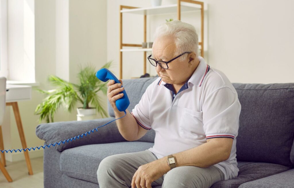 An older adult sitting on the couch and looking at a phone held in his right hand.