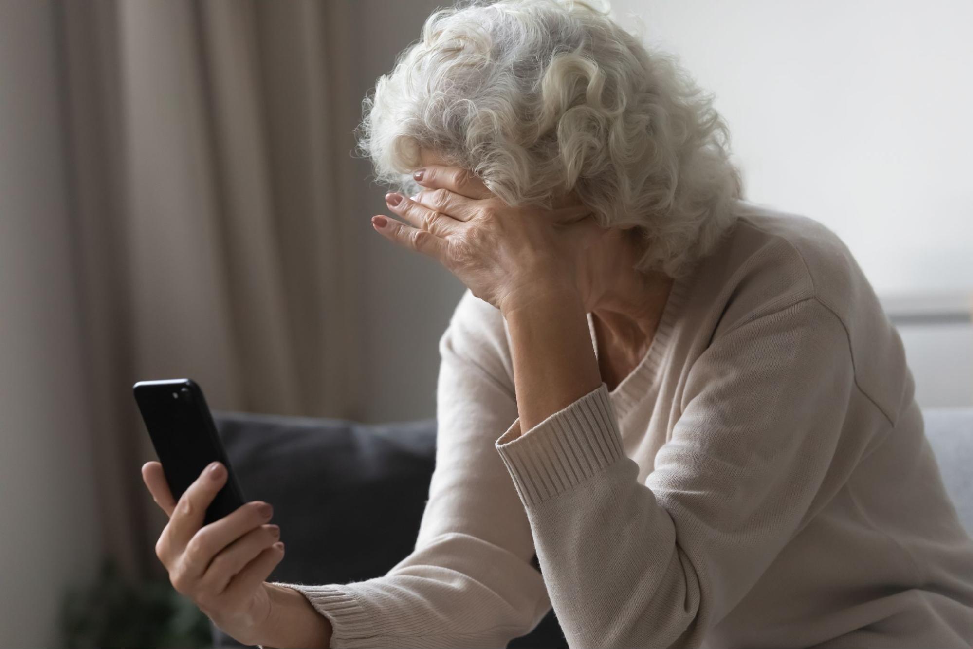 An older adult with dementia sitting on a couch looking at a mobile phone in her right hand while placing her left hand on her forehead.