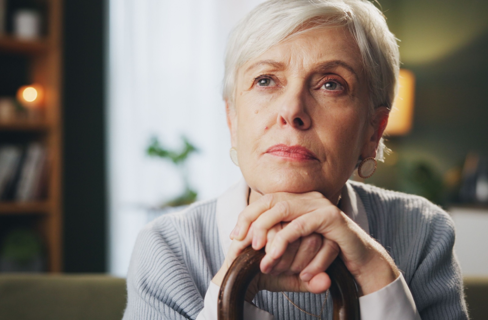 An elderly woman resting her hands and chin on her cane, looking lost in thought.