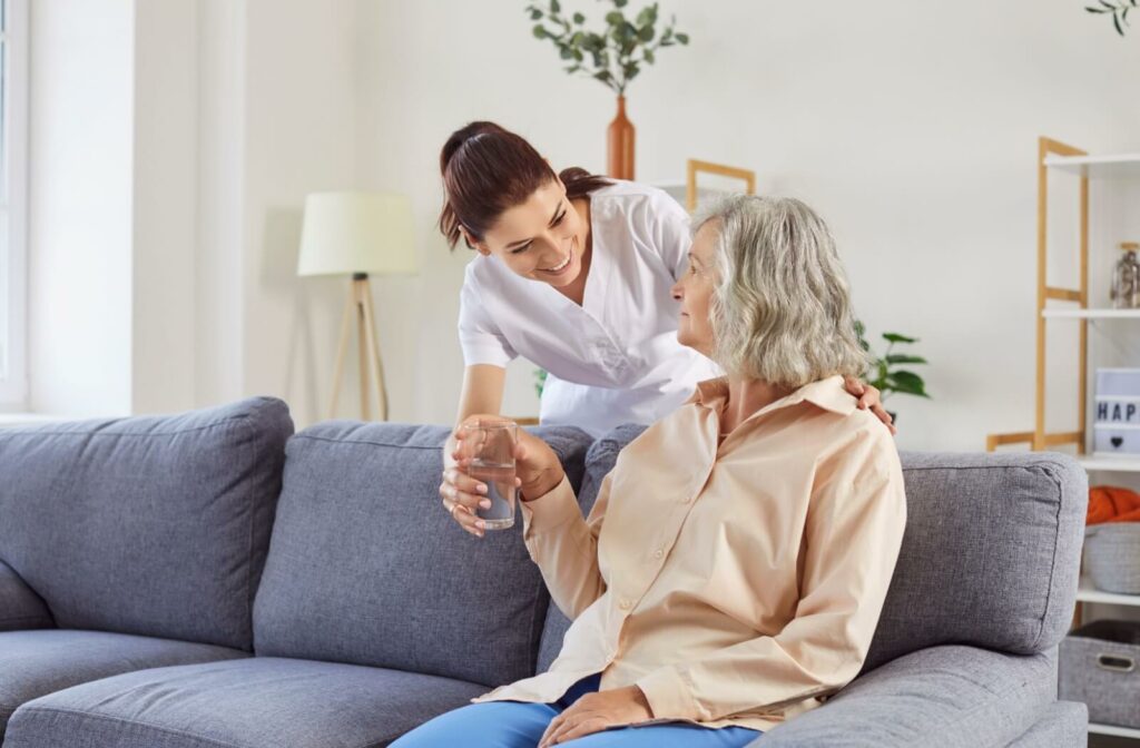 a young aide hands a glass of water to a senior woman sitting on a couch