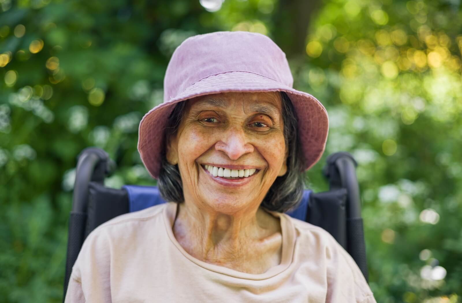 Smiling older adult wearing a pink sun hat and seated outdoors in a shaded green garden area
