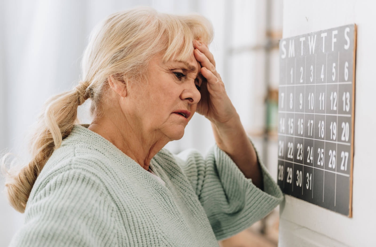 An older adult looks at a wall calendar, expressing stress and confusion.